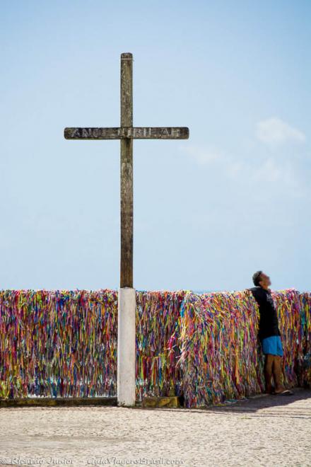 Imagem Crucifixo de madeira e ao fundo fitinhas coloridas amarradas em uma grade.